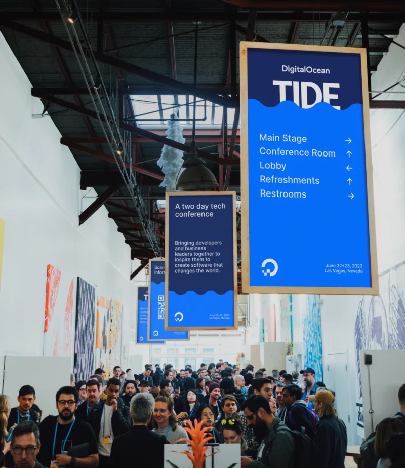 Blue DigitalOcean Tide conference wayfinding signage hanging above a crowded event floor, directing attendees to the main stage and lobby.