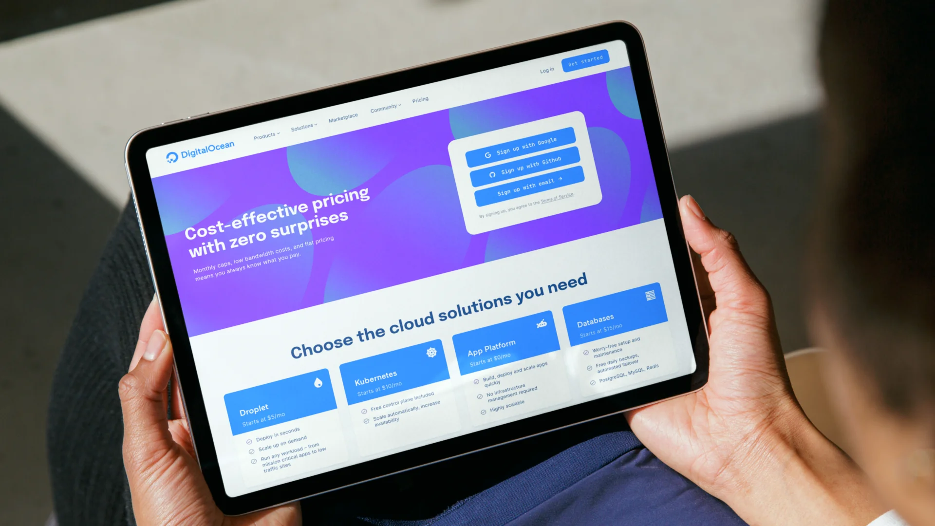 The refreshed DigitalOcean site on a tablet, held by a person, showing a purple 'Cost-effective pricing' hero and a grid of cloud solution cards.